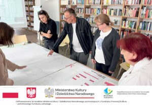 Grupa czterech osób stoi wokół stołu w bibliotece, analizując rozłożone na nim duże arkusze z planami architektonicznymi. Mężczyzna w okularach wskazuje palcem na detal projektu. W tle regały z książkami i ekran projekcyjny. Poniżej belka informacyjna.