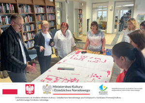 Grupa osób uczestniczy w warsztatach projektowych w przestrzeni bibliotecznej. Stoją przy stole, dyskutując nad rozłożonymi planami z różowymi znacznikami. W tle widoczne regały z książkami oraz przeszklone wejście do innego pomieszczenia. Poniżej belka informacyjna.